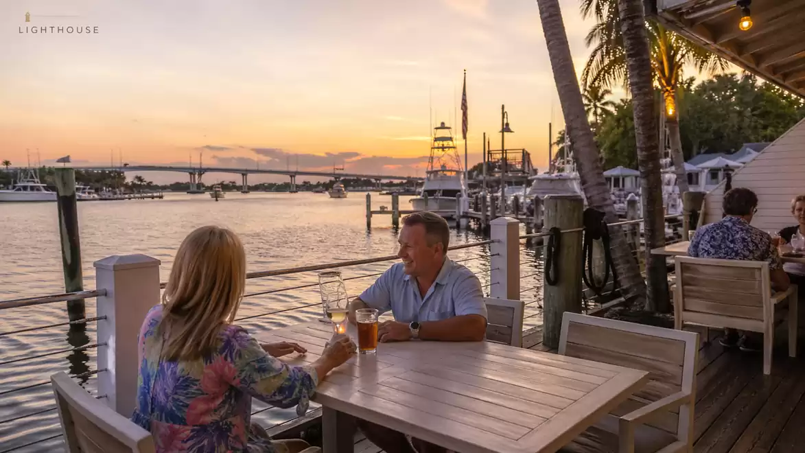 A couple sits at a table in a restaurant overlooking the water at sunset.