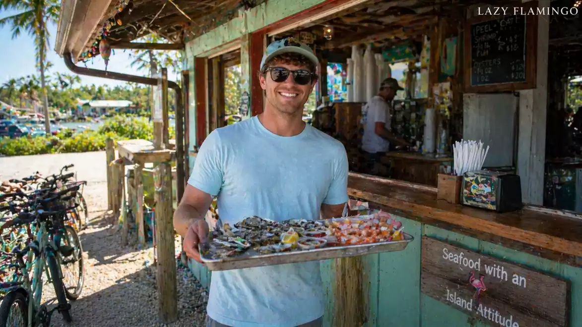 Man in blue clothing carries a tray of seafood in front of a rustic restaurant.