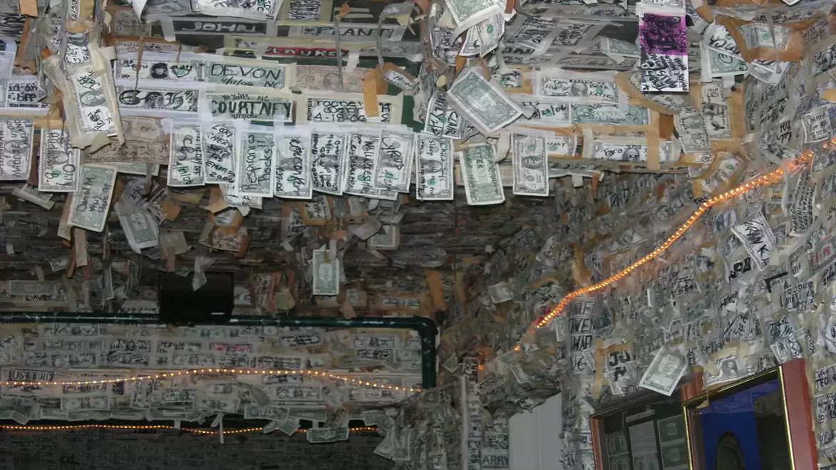 Dollar bills hanging from the ceiling of a bar, covering the walls, with some colorful lights in the background.