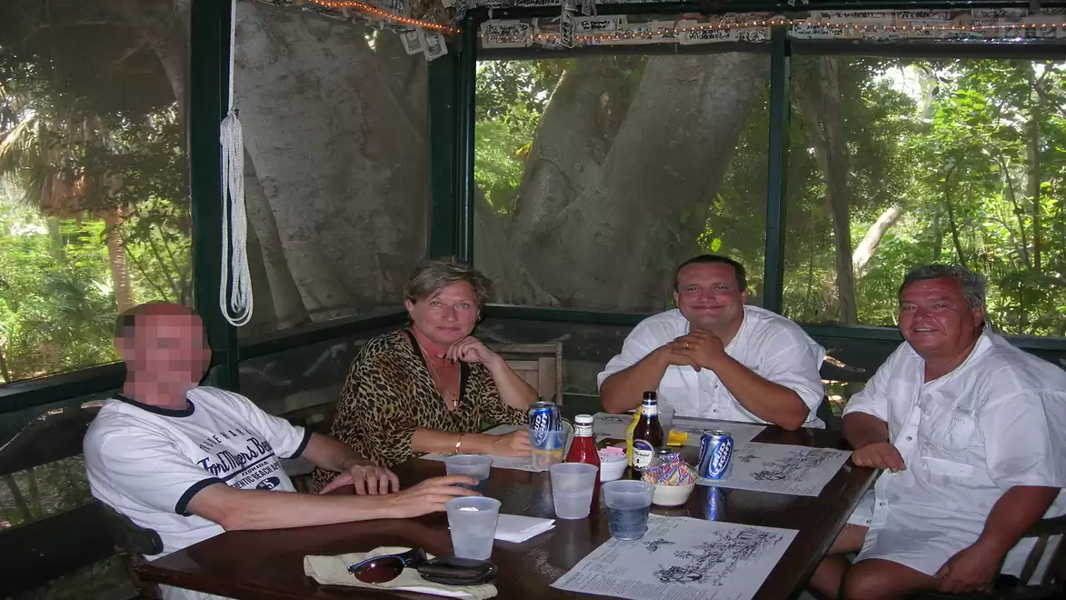 Four people are sitting on a screened porch around a table. Three men and one woman are laughing and relaxing in a casual setting.