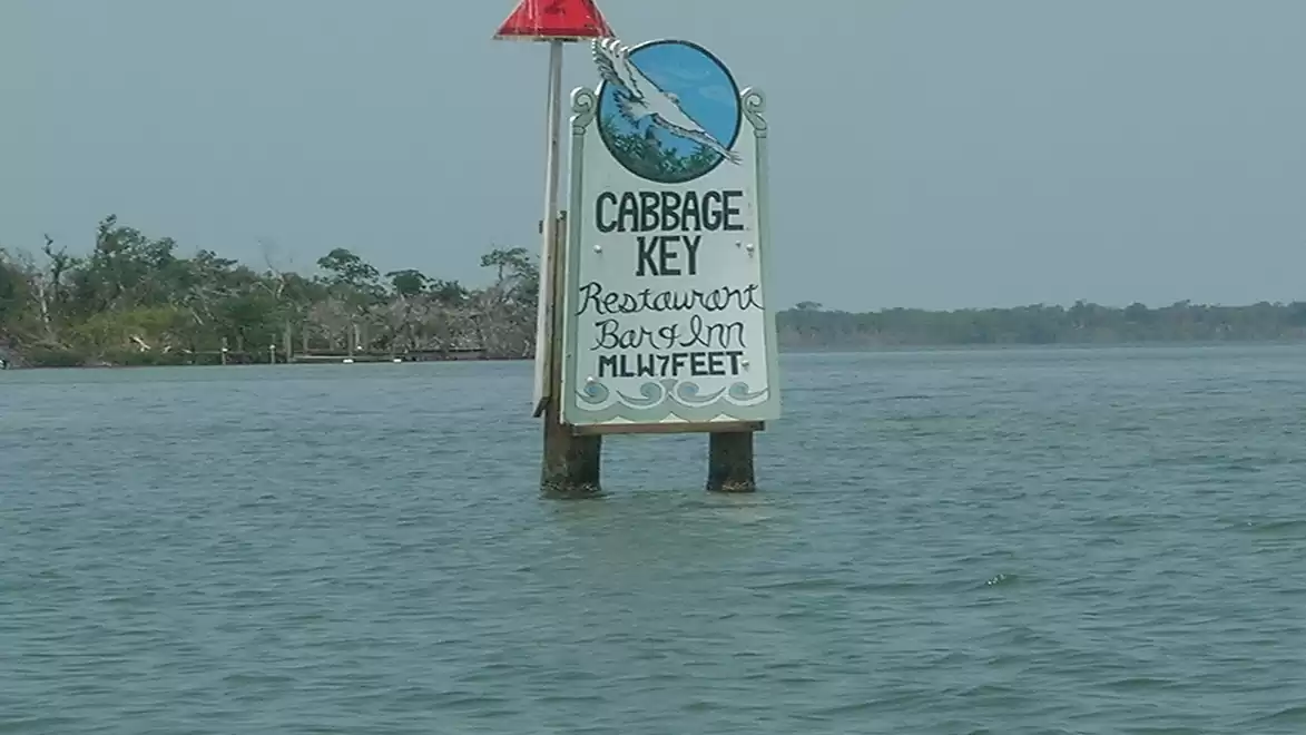 A white sign in the water points the way to Cabbage Key. The sign reads 'CABBAGE KEY Restaurant Bar & Inn 1 1/7 FEET'.