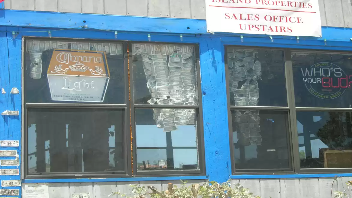 Exterior view of a building with blue window frames, a neon beer sign, and a sign indicating a sales office upstairs.
