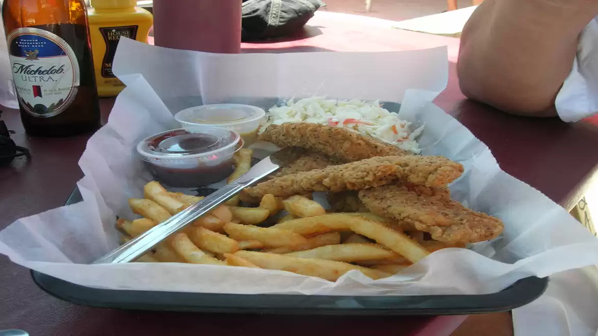 A plate with fries, fried chicken, and coleslaw on an outdoor table. Next to it are bottles of sauces.