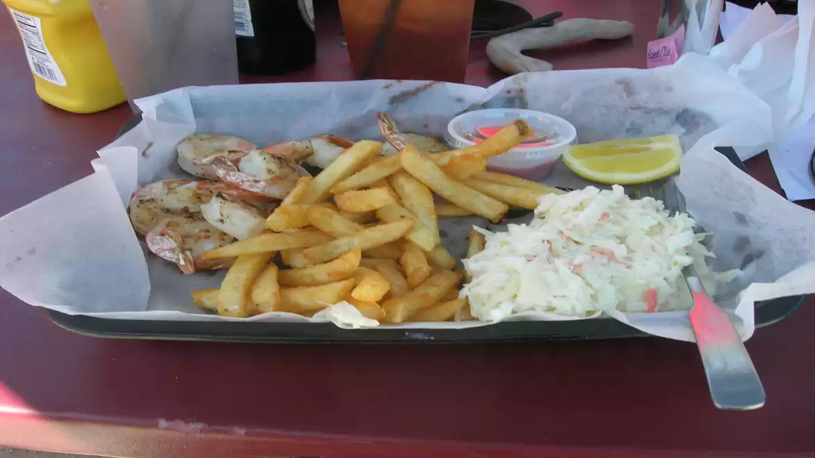 A tray with grilled shrimp, French fries, coleslaw, and a lemon wedge on a table.