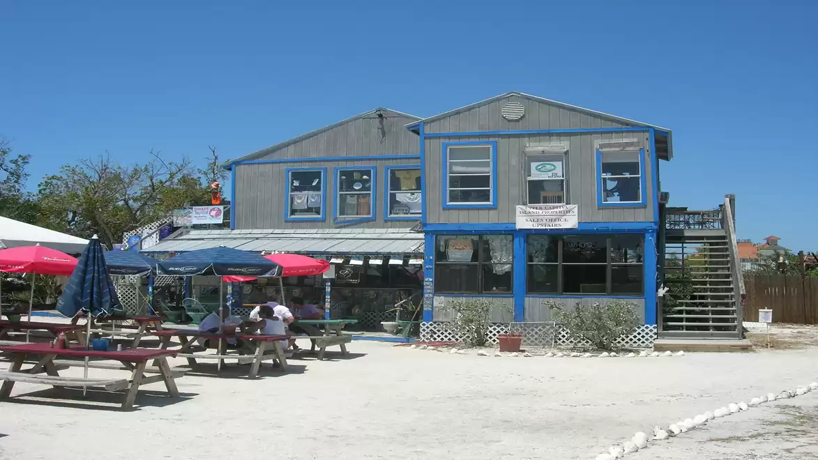 A two-story building with blue trim and several windows. In front, there are tables with umbrellas in the open air. The sky is clear and blue.