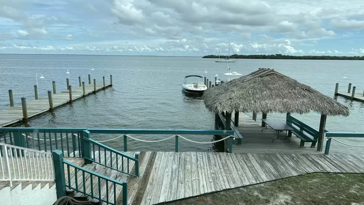 A boat dock with a thatched-roof pavilion on calm waters. In the background, clouds are visible in the sky and smaller boats are on the water.