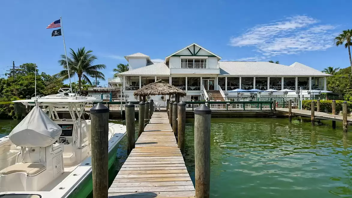 An idyllic coastal restaurant with a long wooden pier, boats, and palm trees under a bright blue sky.