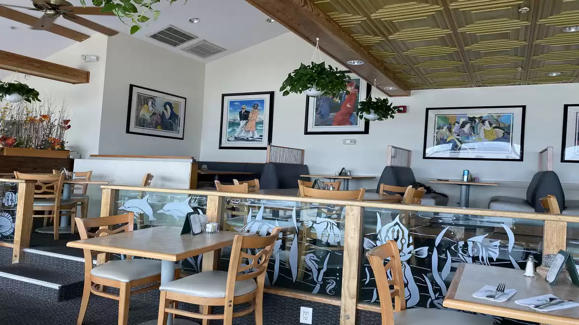 A modern restaurant with wooden decor, ceiling plants, and artworks on the walls. The space appears calm and inviting with set tables and cushioned seating.