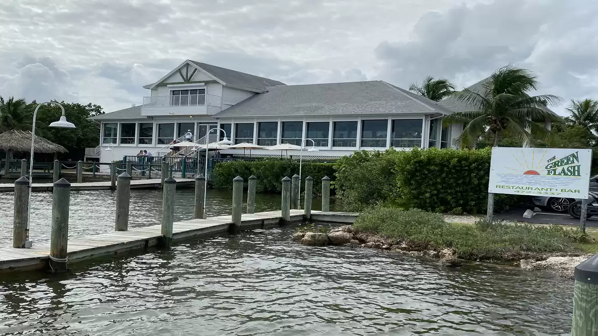 A restaurant with a gray roof on a riverbank, surrounded by palm trees, with a sign reading 'Green Flash'.