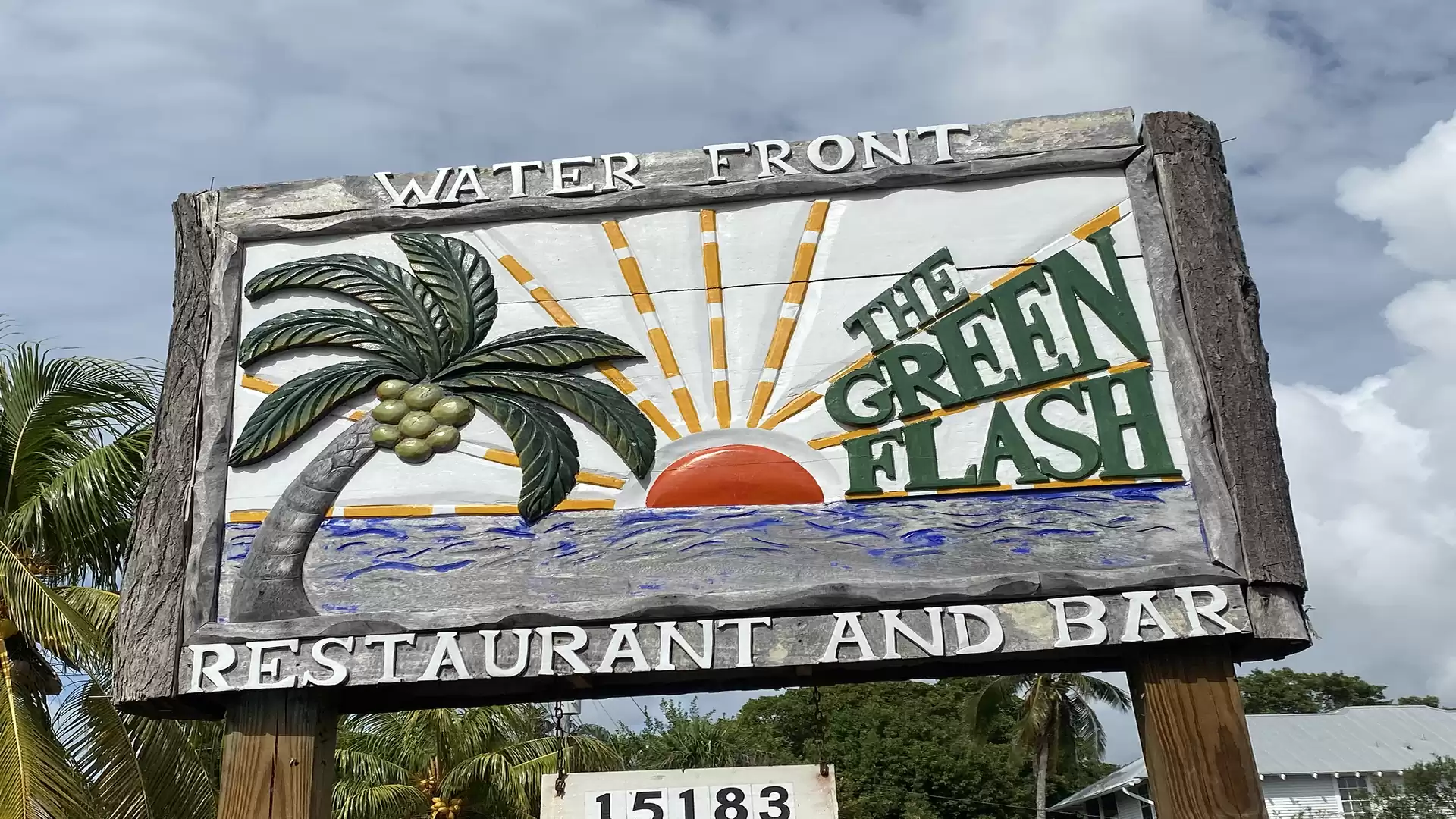 A large wooden sign reading 'The Green Flash', featuring a palm tree, a setting sun, and the sea. It is located at a restaurant and bar called Waterfront.