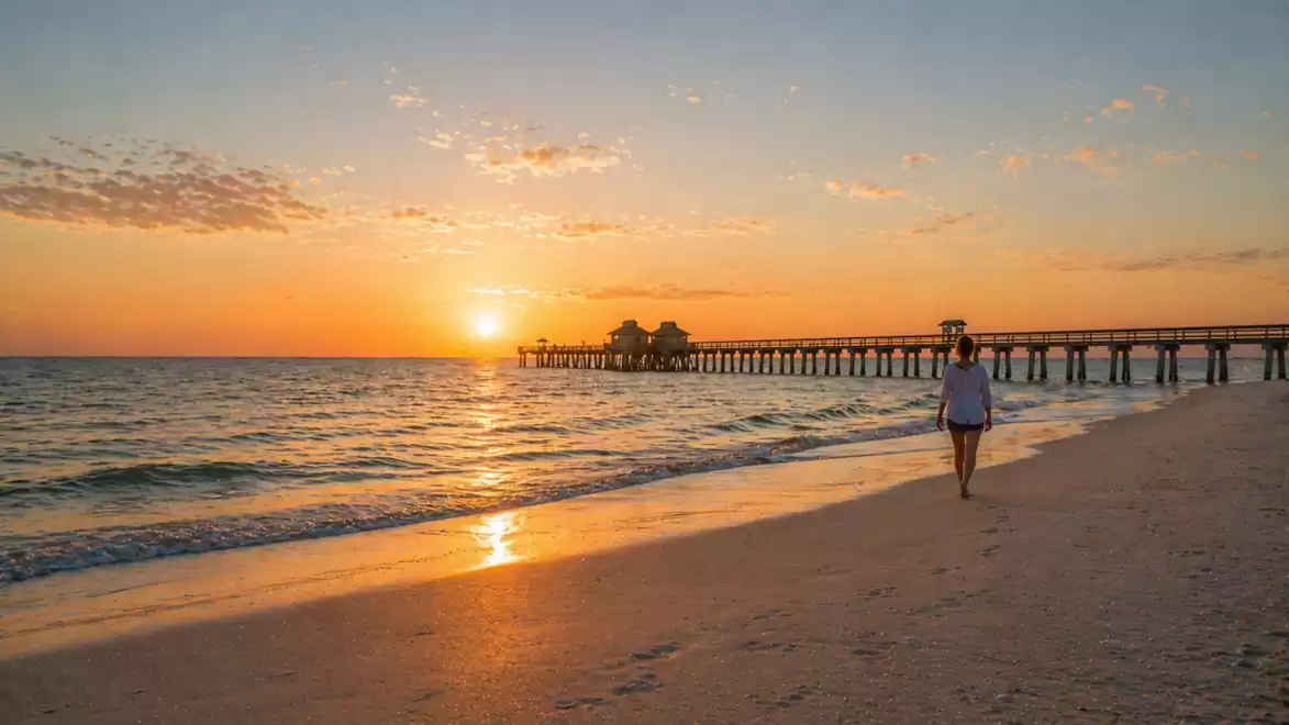 A sunset on the beach with a person walking along the water. In the background, a long pier extends into the sea.