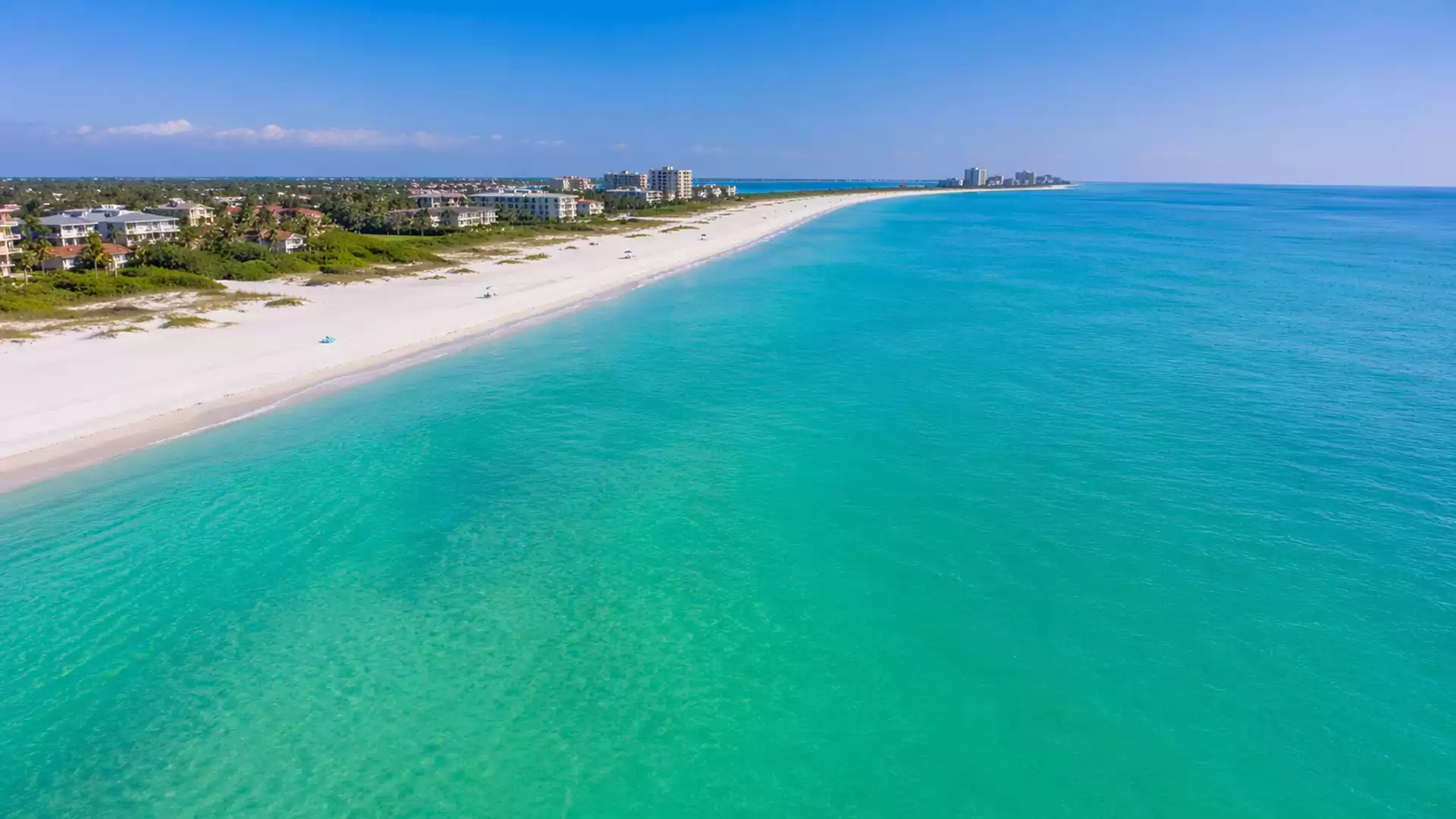 Aerial view of a sunny tropical beach with clear turquoise water and white sand, bordered by lush vegetation and scattered buildings on the horizon.