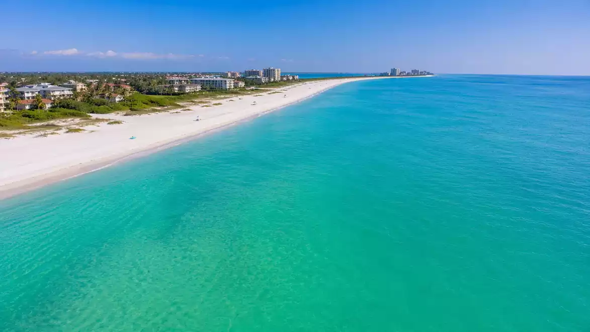 Aerial view of a sunny tropical beach with clear turquoise water and white sand, bordered by lush vegetation and scattered buildings on the horizon.