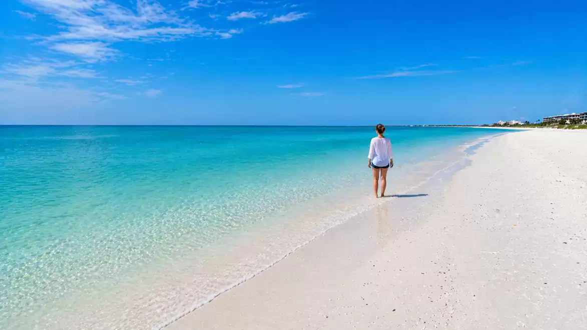 Woman walking on a sunny sandy beach with clear turquoise water under a blue sky.