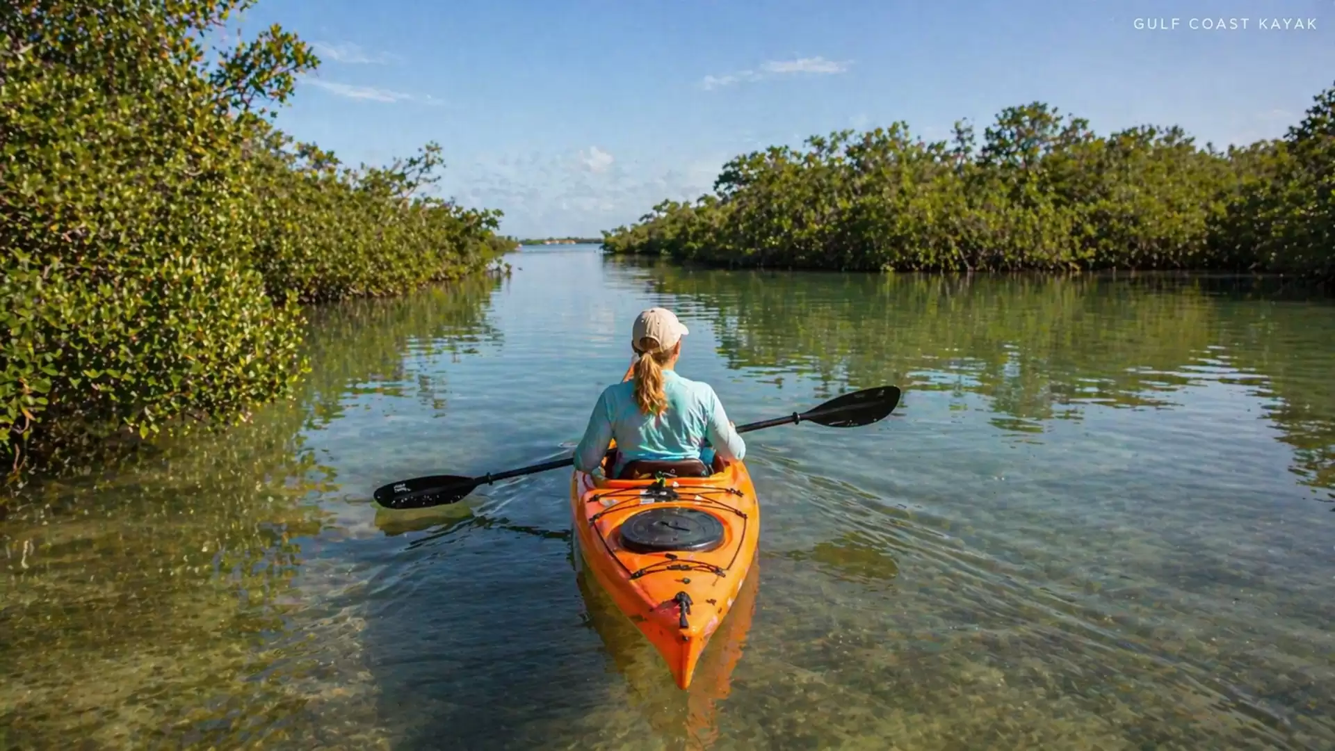 Person in an orange kayak paddling through calm waters, surrounded by green mangroves under a clear sky.