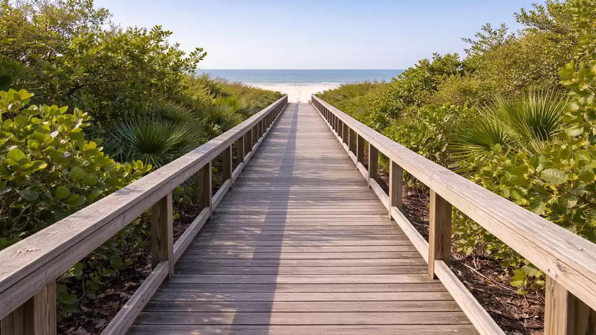 A wooden boardwalk leads through lush green vegetation to a bright sandy beach with a blue sky in the background.