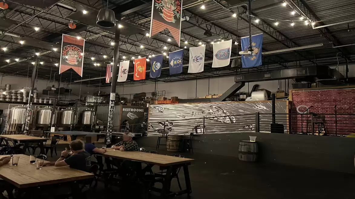 A brewery hall with wooden tables, hanging banners, and large brewing tanks under an illuminated ceiling.
