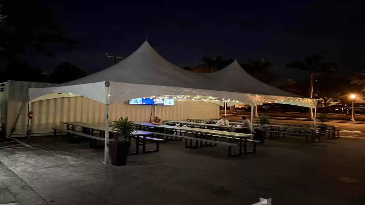A night-time outdoor area with a large white tent covering tables and benches. Some people are sitting at the tables. String lights decorate the tent.