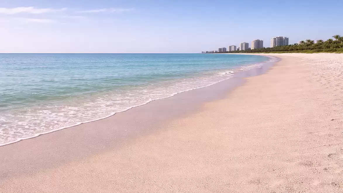 A serene sandy beach with clear blue water and buildings on the horizon.