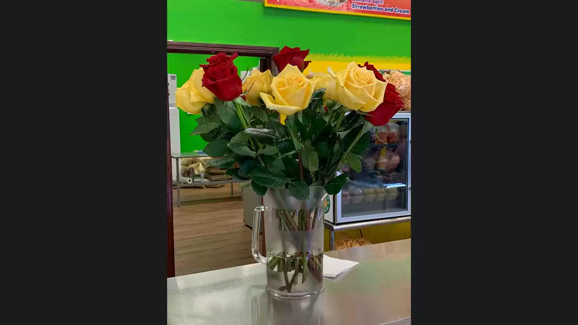A bouquet of red and yellow roses in a vase on a table. In the background, a green painted wall and a storefront are visible.