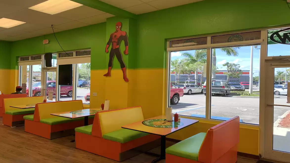 Interior view of a colorful restaurant with green and yellow walls, one of which is decorated with a large mural of a superhero. There are several orange-yellow booth seats. Large windows provide views of the street and parked cars.