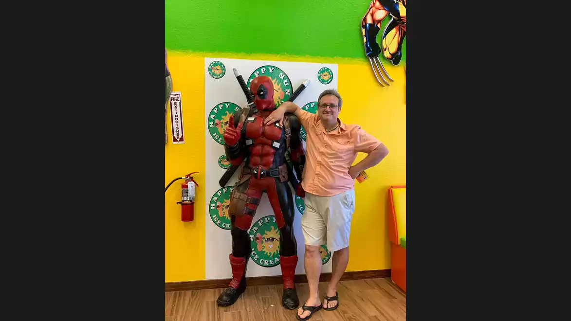 Man posing next to a superhero figure in a colorful room with green and yellow walls.