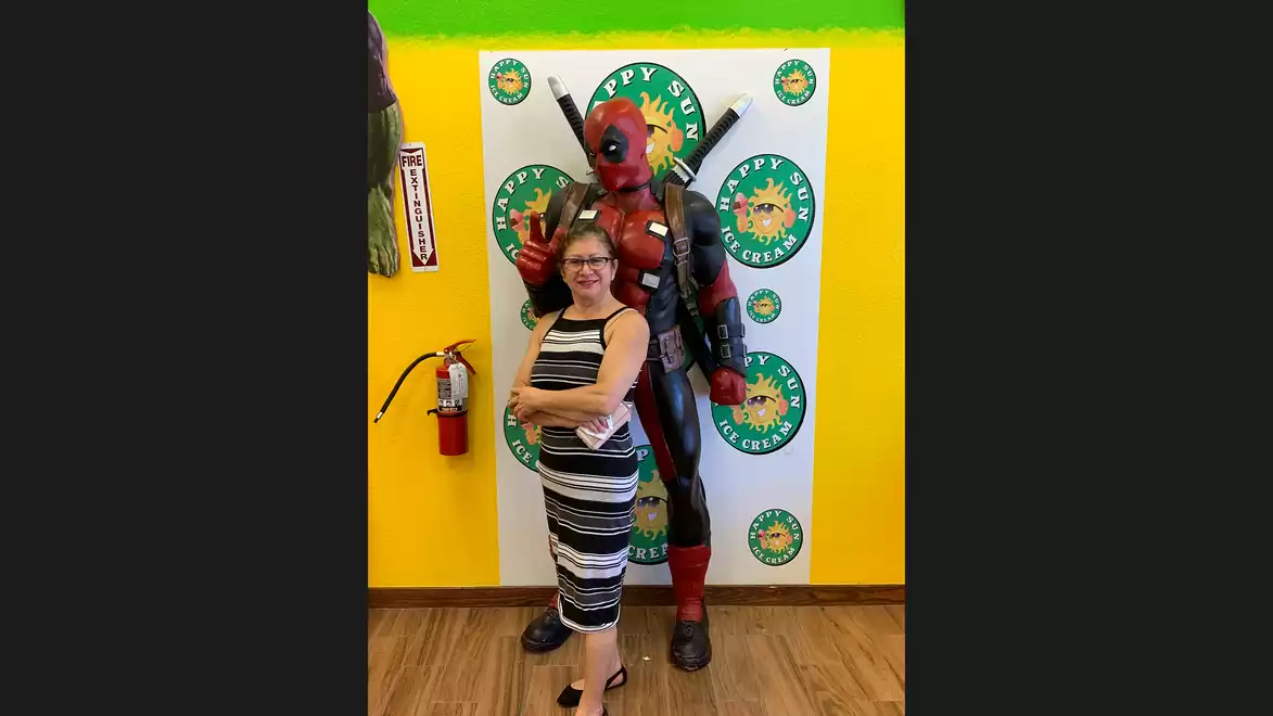 A woman stands smiling in front of a poster with a person dressed as a superhero posing. The room is colorfully and cheerfully decorated.