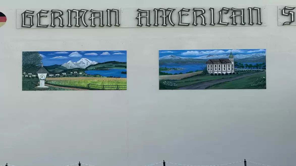 Mural on a building with the inscription 'German American', displaying two landscape paintings: a hilly landscape with a lake and a large manor house.