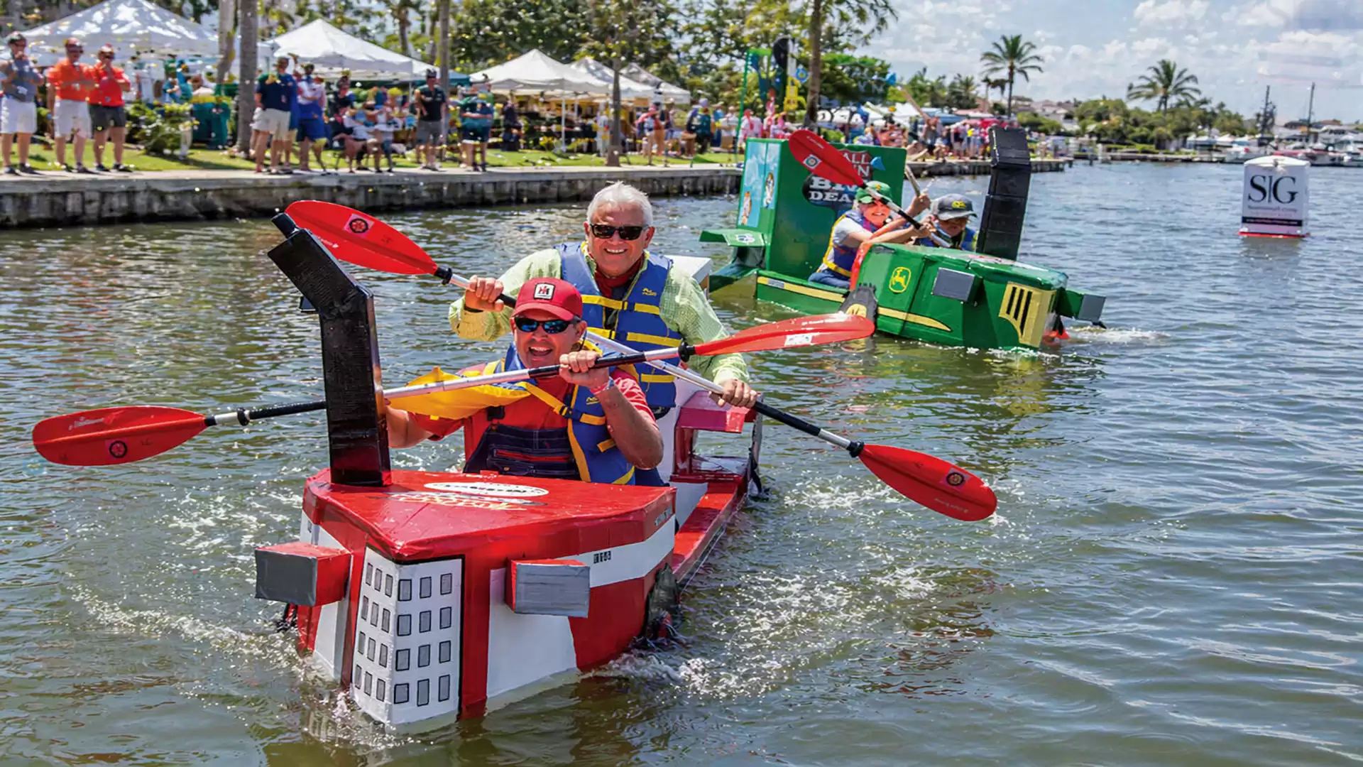 Participants in a creative boat race paddle in colorful, homemade cardboard boats on a canal.