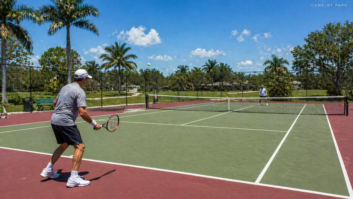 Two men playing tennis on an outdoor court on a sunny day, surrounded by palm trees.