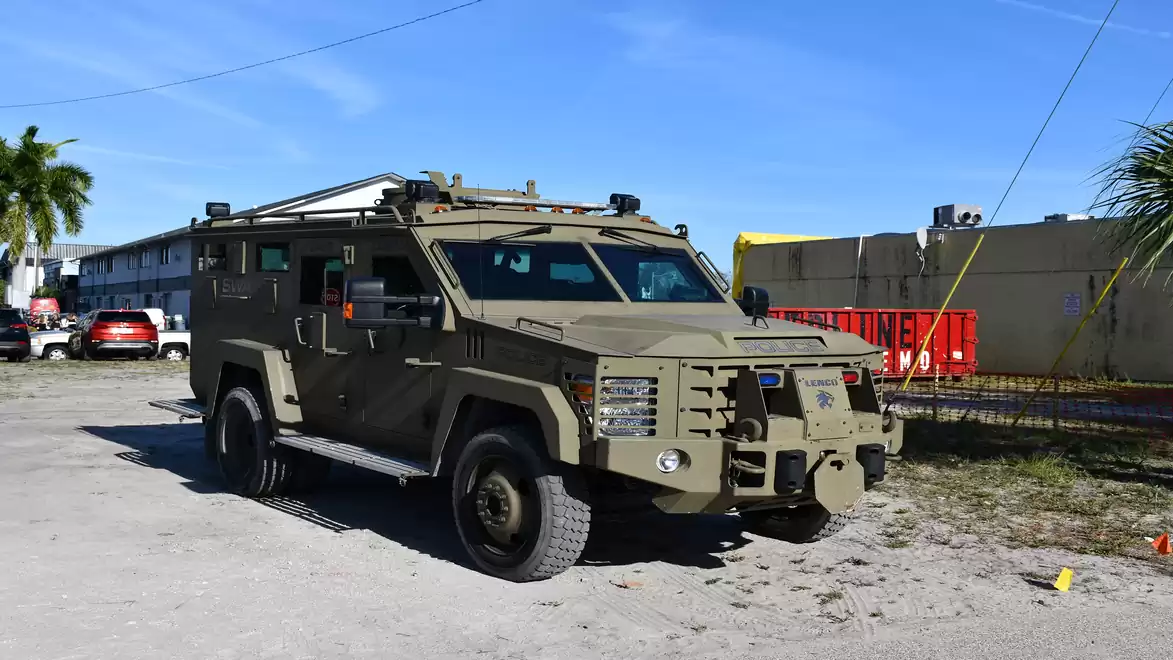 An armored vehicle is parked outdoors, surrounded by palm trees and modern buildings in sunny weather.