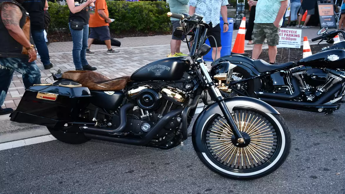 A shiny black motorcycle with gold-spoked wheels stands on a street, surrounded by people at an event.