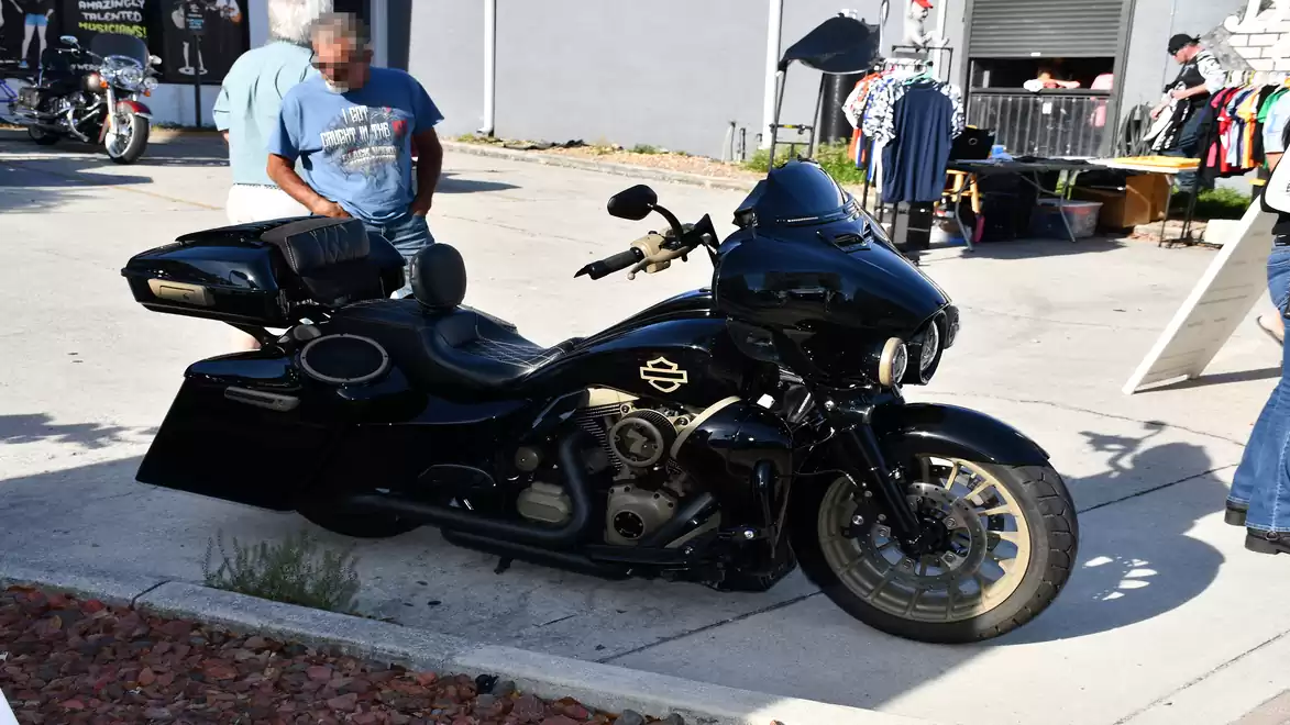 A black motorcycle is parked on a sidewalk, with a man in a blue t-shirt standing next to it. Market stalls are visible in the background.