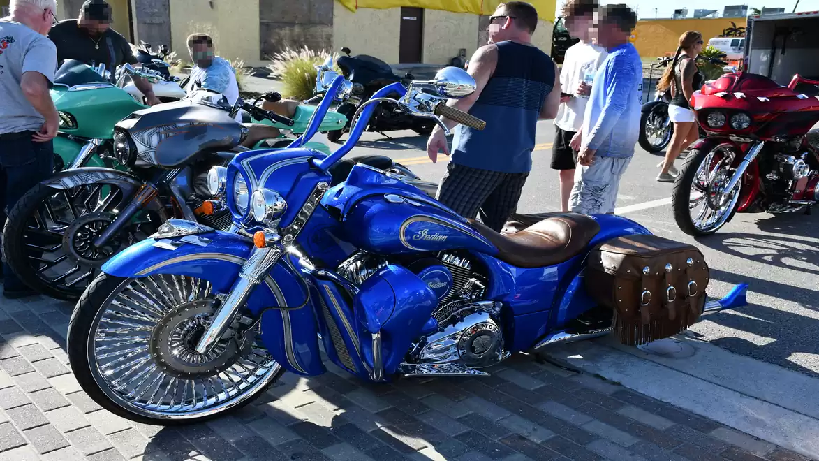 Blue custom motorcycles with spoked wheels and brown leather bags are parked side by side at a motorcycle exhibition. In the background, people can be seen admiring the motorcycles.