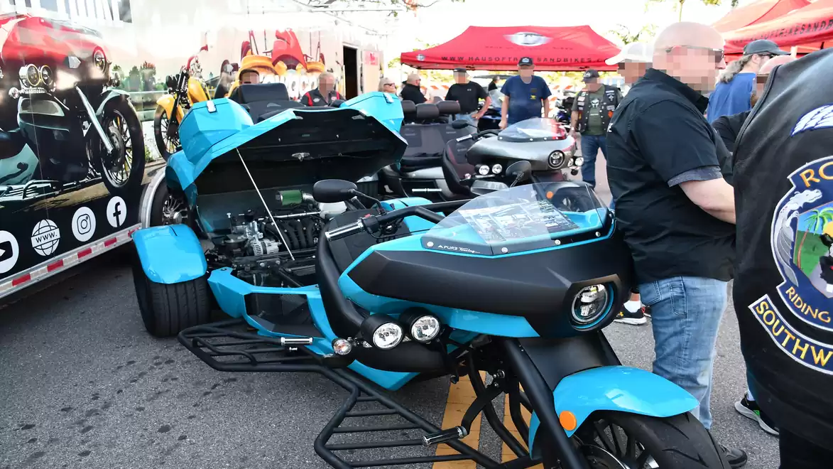 A blue three-wheeled motorcycle with an open hood is parked on a busy street during a motorcycle exhibition. People surround the motorcycle, and red tents are visible in the background.