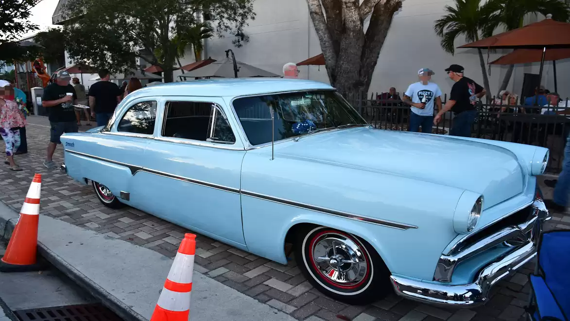 A classic light blue car is parked on a cobblestone street. People and trees are visible in the background.