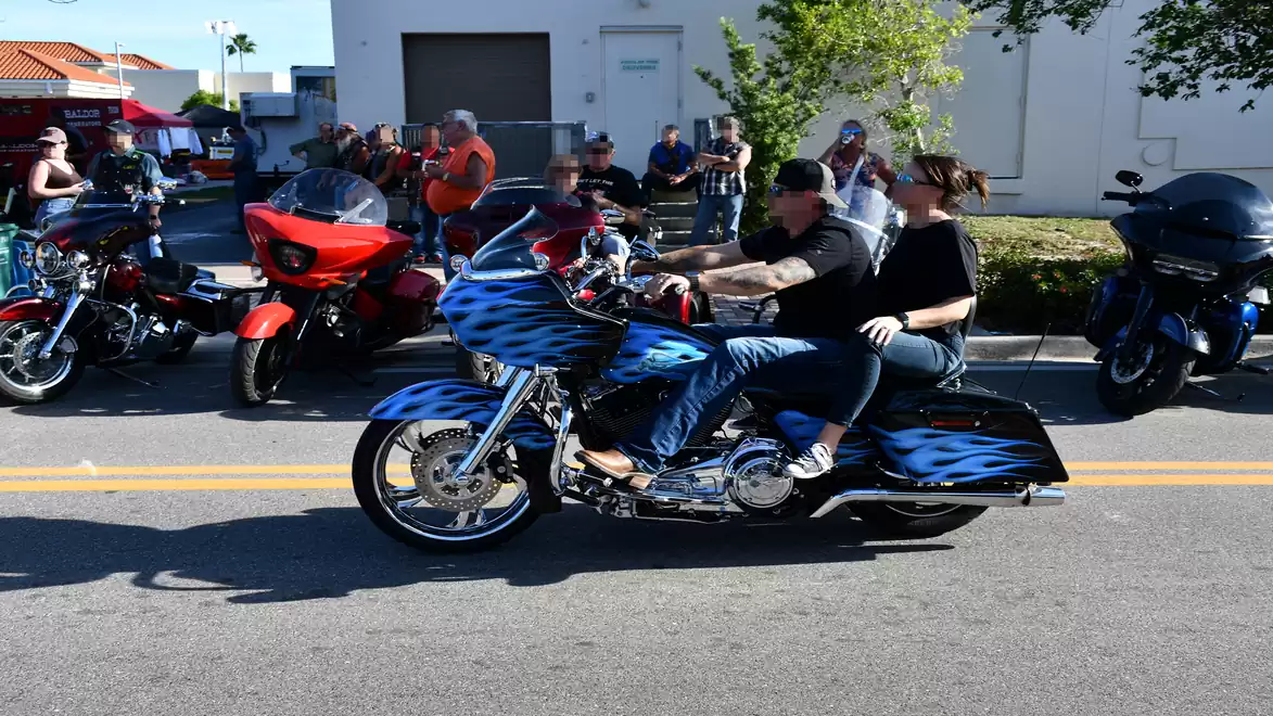 A couple rides a motorcycle with a blue flame paint job at a bike rally, surrounded by other motorcycles and spectators.