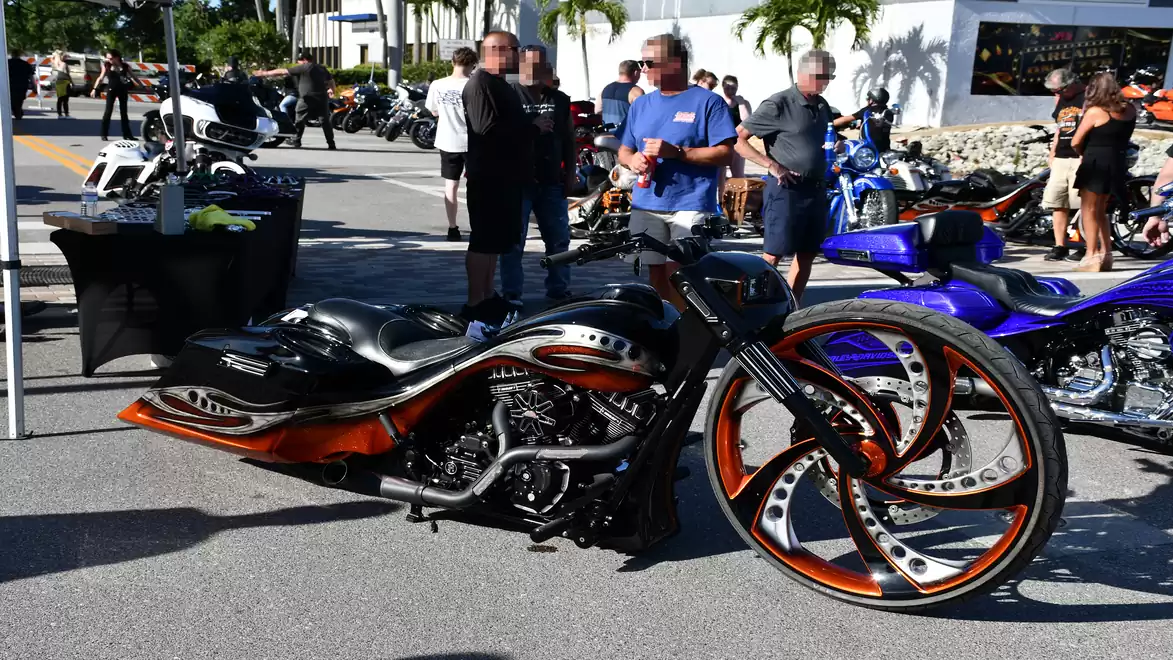 A stylish custom motorcycle with striking orange rims is on display at an outdoor motorcycle exhibition, surrounded by visitors admiring it.