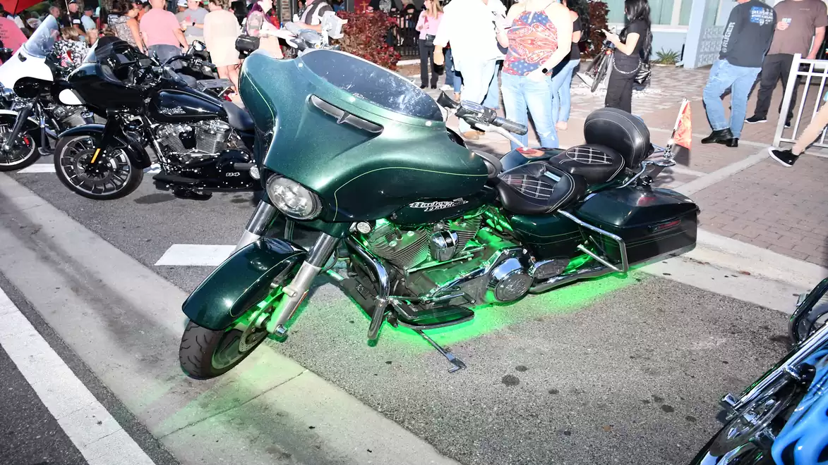 A green motorcycle with glowing underbody lighting stands on a street, surrounded by people and other motorcycles.
