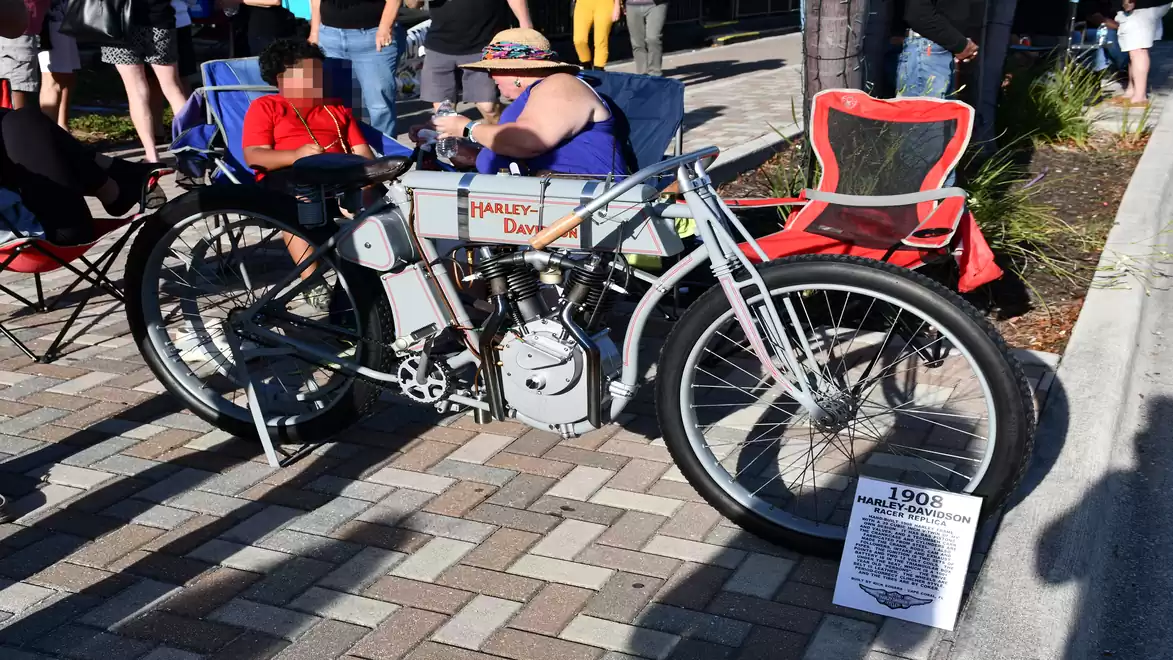 An old Harley-Davidson motorcycle from 1908 is displayed on a paved street, surrounded by several people admiring the historic vehicle. Seating is also available.