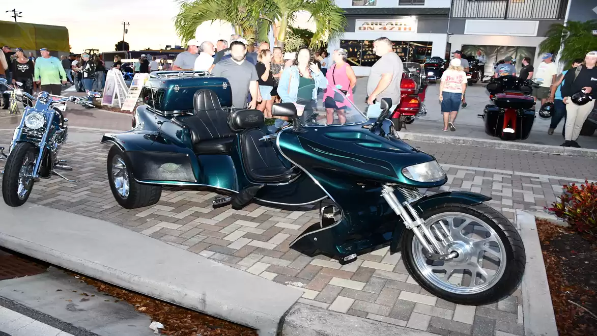 An impressive trike is parked on a paved street at an event, surrounded by people and additional motorcycles.