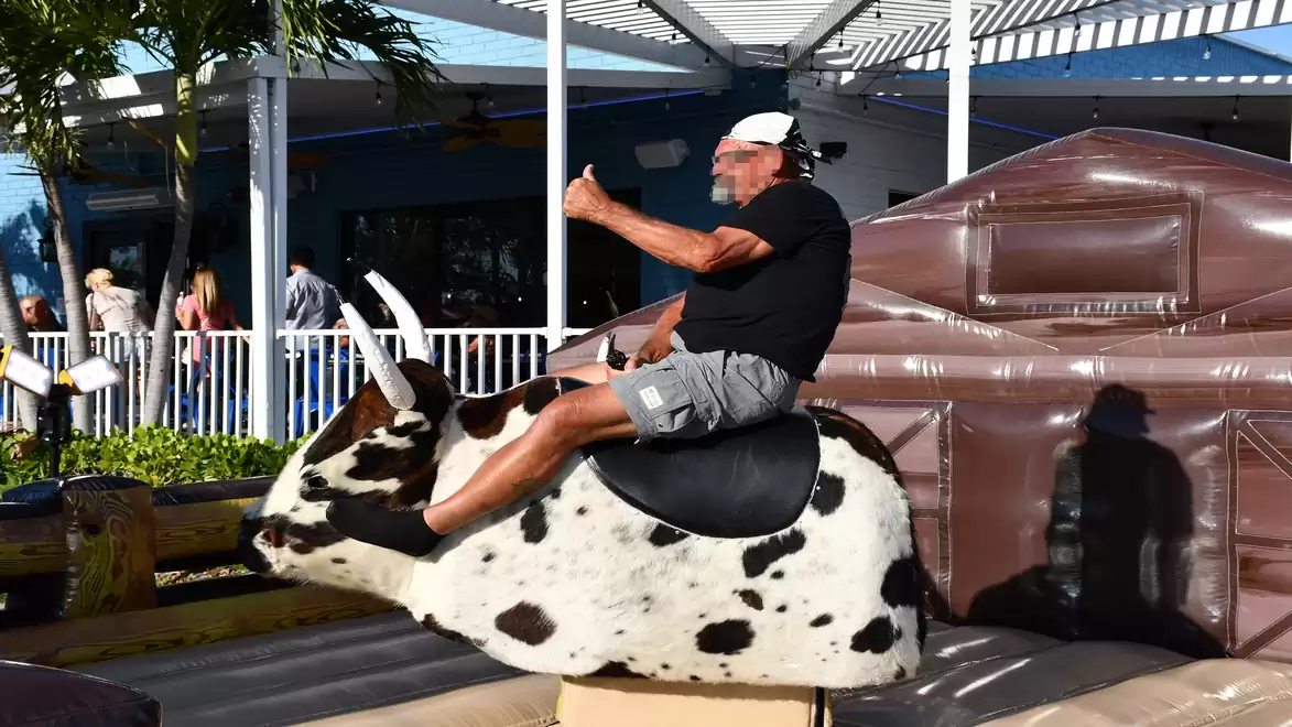A man in shorts and a cap sits on a mechanical bull, giving a thumbs up. In the background, a patio and people are visible.