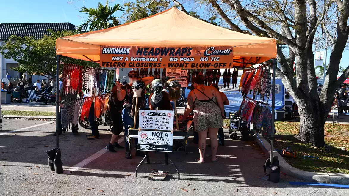 A market stall with an orange tent selling handmade headwraps. Several people are standing under the tent, looking at the products, surrounded by colorful wraps.
