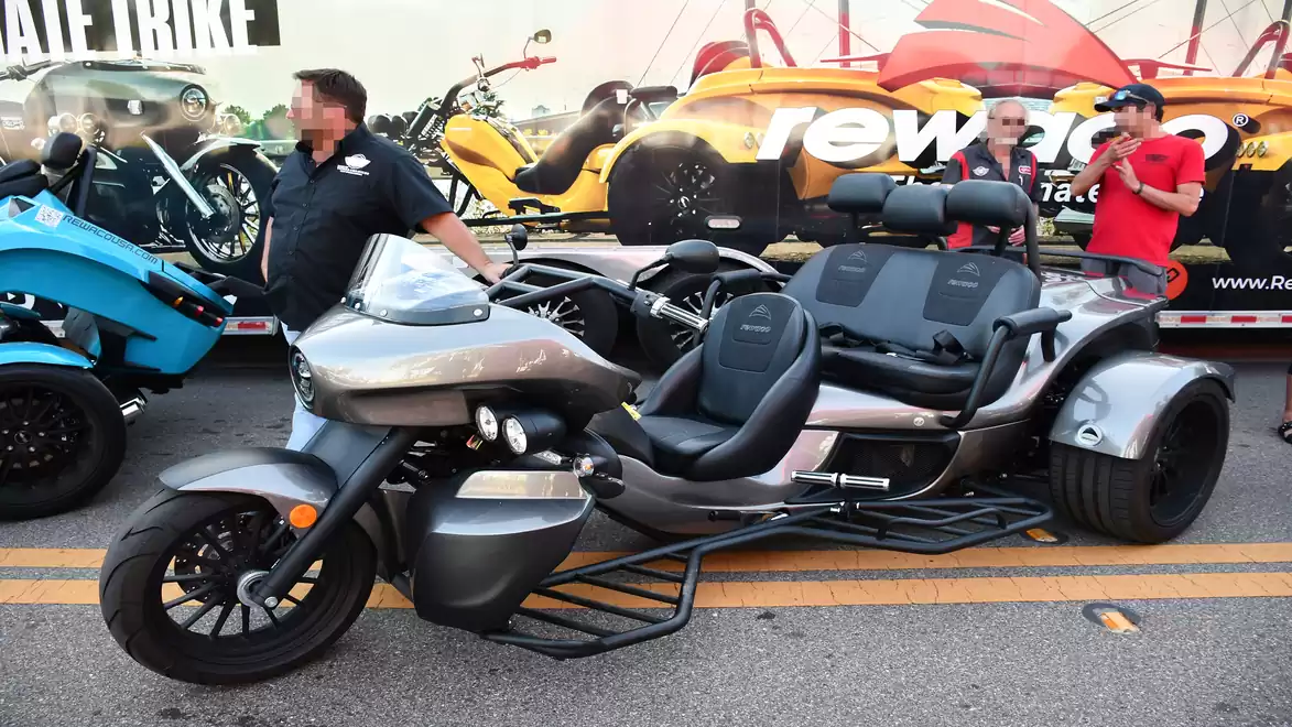 A futuristic-looking, three-wheeled motorcycle is showcased on a street next to a man. In the background, there is a trailer with red and yellow graphics and two other people.