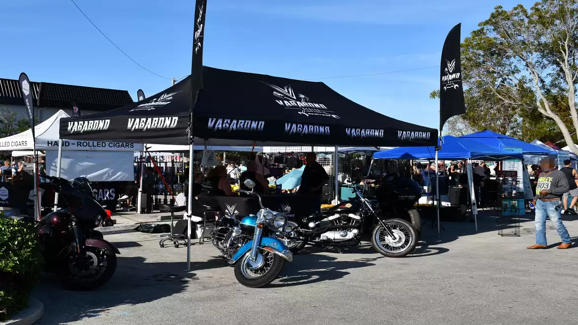 Motorbike event with tents and exhibitors under a bright blue sky. Several motorcycles are parked in front of the tents.
