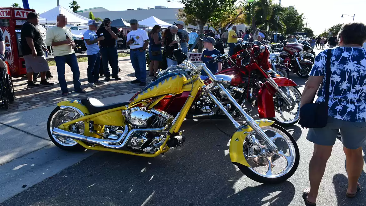 Several motorcycles are lined up at a gathering on a street. People are looking at the displayed bikes. A brightly colored yellow motorcycle is prominently visible in the foreground.