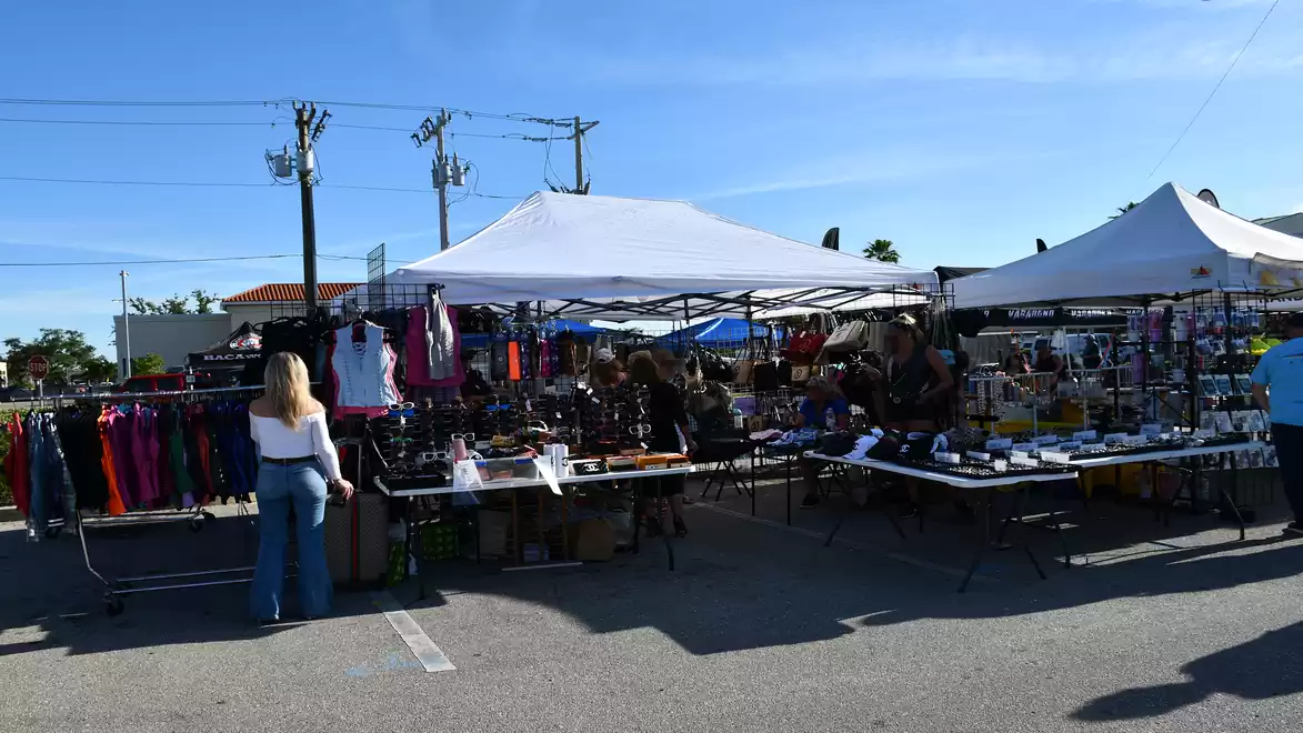 An outdoor market with several stalls selling clothes and other items. People are browsing and shopping. The sky is clear and blue.