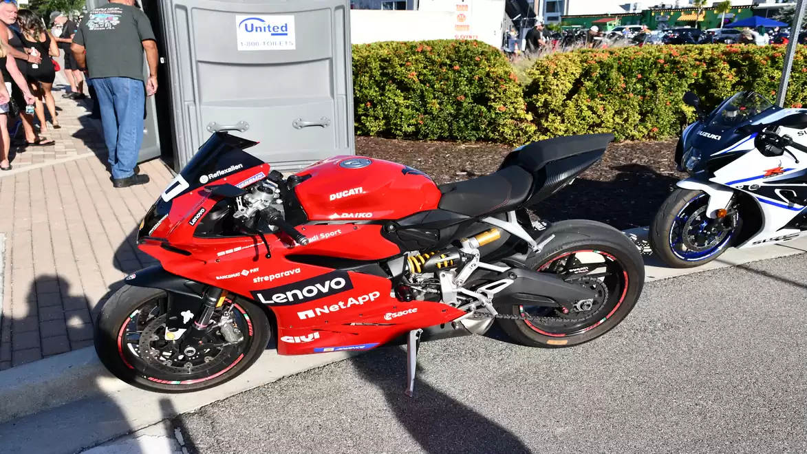 A red sports motorcycle with Lenovo and NetApp stickers is parked next to a blue and white motorcycle in a parking lot in front of a row of bushes and a portable toilet cabin.