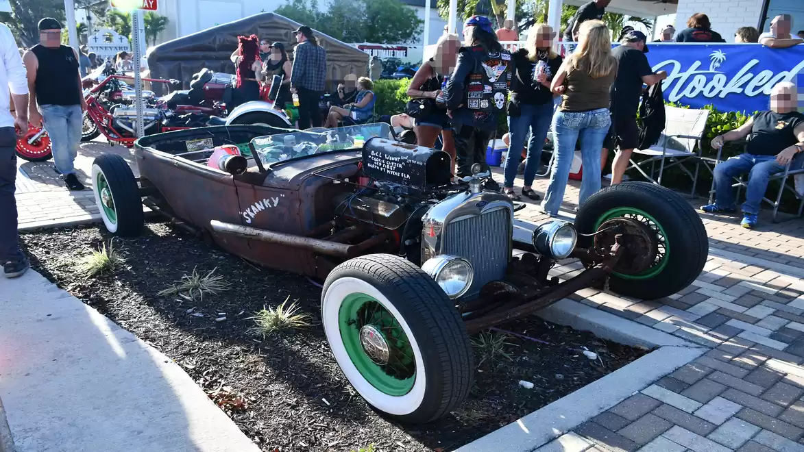 A rustic Hot Rod with an exposed engine and green rims is parked on a pedestrian walkway, surrounded by people at a car show.