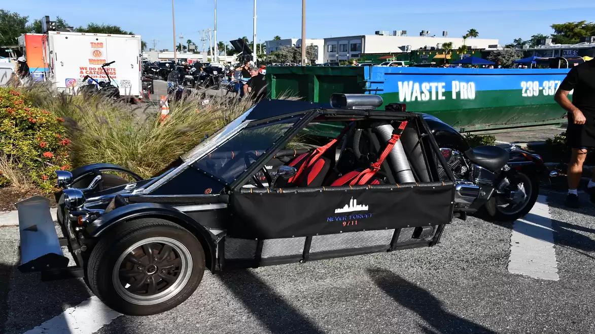 A three-wheeled vehicle with a black canopy and red seatbelts parked next to a dumpster outdoors. Cars, plants, and buildings are visible in the background.